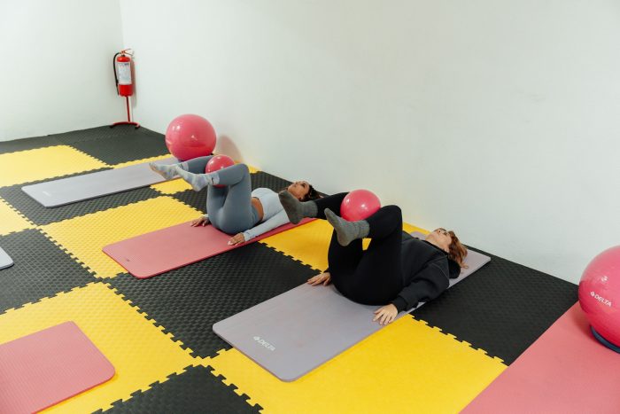 Two women lying on their backs on yoga mats with fitness balls between their knees