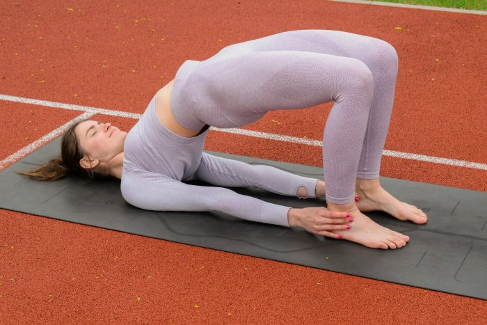 Woman wearing a purple gym set whilst stretching on a mat with her hips in the air whilst laid on her back and touching her ankles